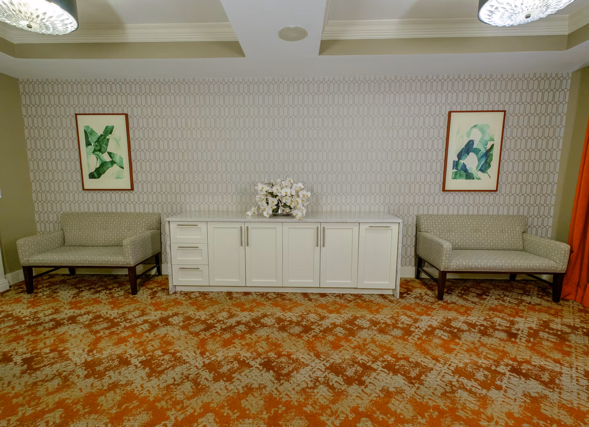 A seating area with two beige upholstered chairs on either side of a white cabinet with drawers and doors. The wall behind has a geometric patterned wallpaper with two framed abstract green artworks. A floral arrangement is placed on top of the cabinet. The floor is covered with an orange and beige patterned carpet.