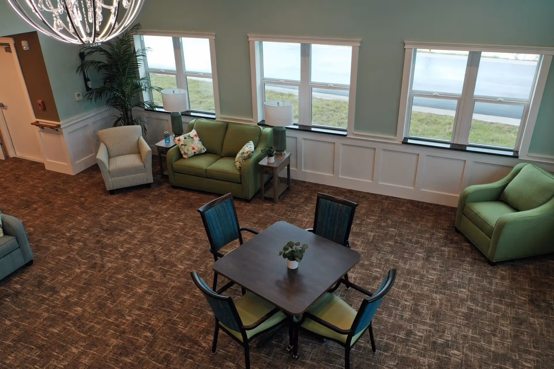 A bright and spacious living room area with large windows letting in natural light. The room features a green loveseat with floral pillows, two green armchairs, a small table with a plant in the center surrounded by four chairs, and two side tables with lamps. There is a large decorative chandelier hanging from the ceiling and a tall potted plant in the corner.