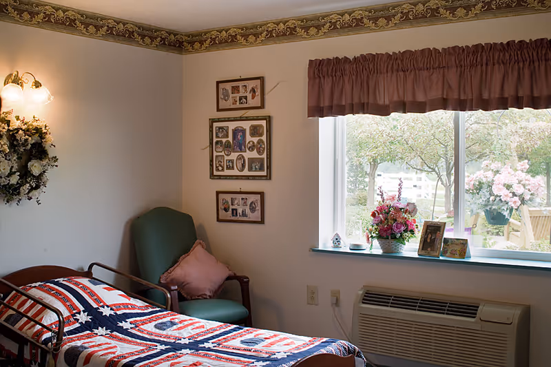 A cozy senior bedroom with a bed covered by a patriotic quilt, a green armchair, framed photos on the wall, and a window sill with flowers and picture frames.