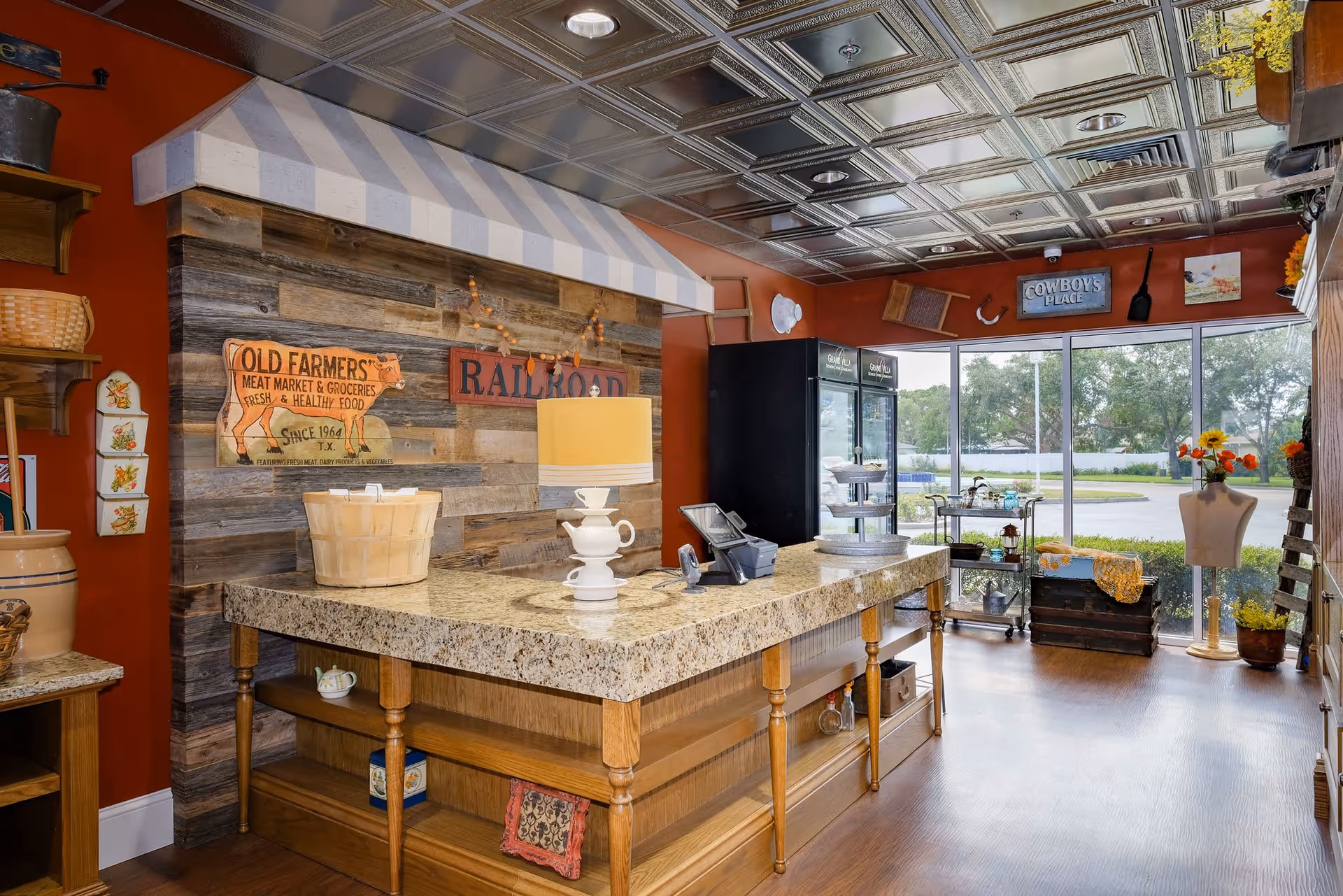 Interior of a rustic-themed room with a large granite countertop island in the center, wooden shelves with decorative items, a striped awning above a wooden wall with vintage signs, a lamp on the counter, and large windows showing greenery outside.