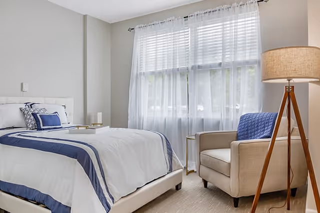 A bright bedroom featuring a neatly made bed with white and blue bedding, a beige armchair with a blue cushion, a small round side table, and a tall floor lamp with a beige shade. The room has a large window with sheer white curtains allowing natural light to fill the space.