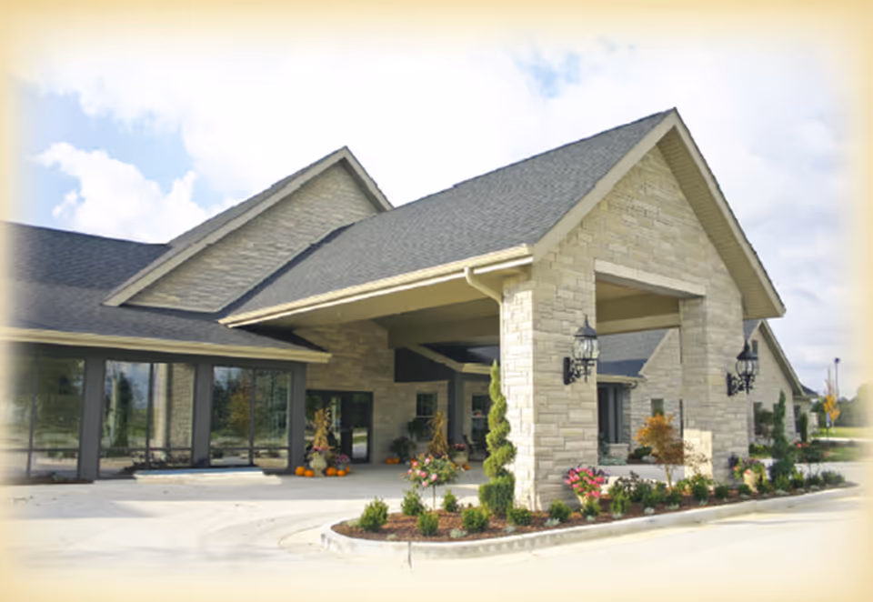 Front entrance of a stone-clad senior living facility with a covered porte-cochere, drive-up loop, and landscaped flowerbeds.