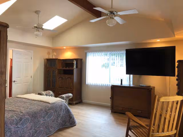 Spacious bedroom featuring a bed with a patterned coverlet, wooden cabinets and rocking chair, a wall-mounted TV, ceiling fans and skylights, and a large window with vertical blinds.