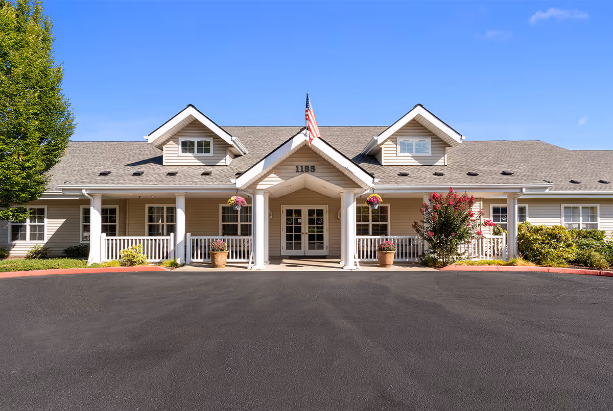 Front exterior view of a single-story senior living facility building with beige siding, white columns, and a covered entrance. There are hanging flower pots and potted plants near the entrance, an American flag above the door, and a clear blue sky in the background.