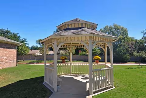 Wooden gazebo in a grassy courtyard with a brick building nearby and potted plants.