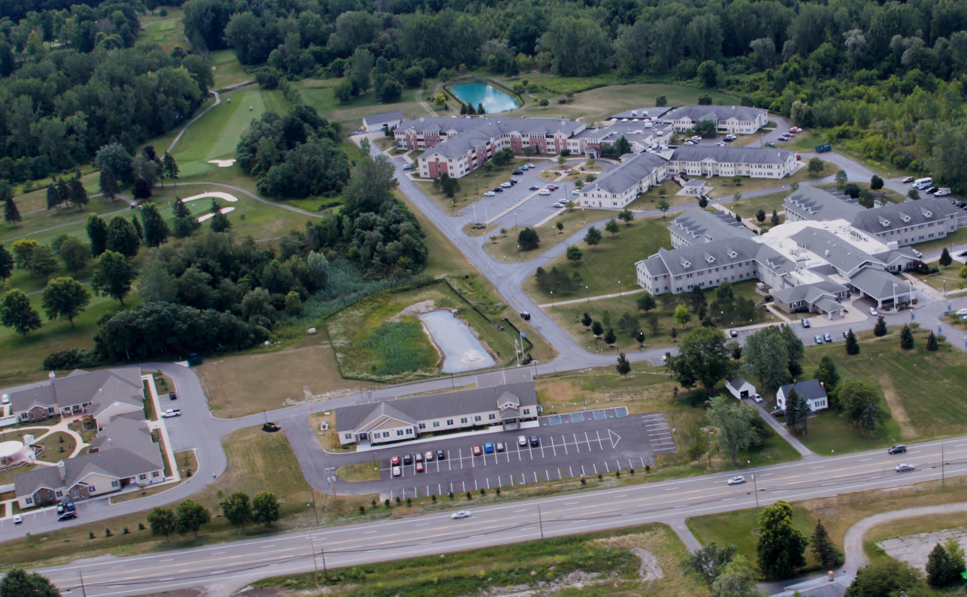 Aerial view of The GreenFields Continuing Care Community showing multiple large buildings, parking lots, a small pond, surrounding trees, and nearby roads. The area includes green spaces and appears to be a well-maintained campus set in a semi-rural environment.