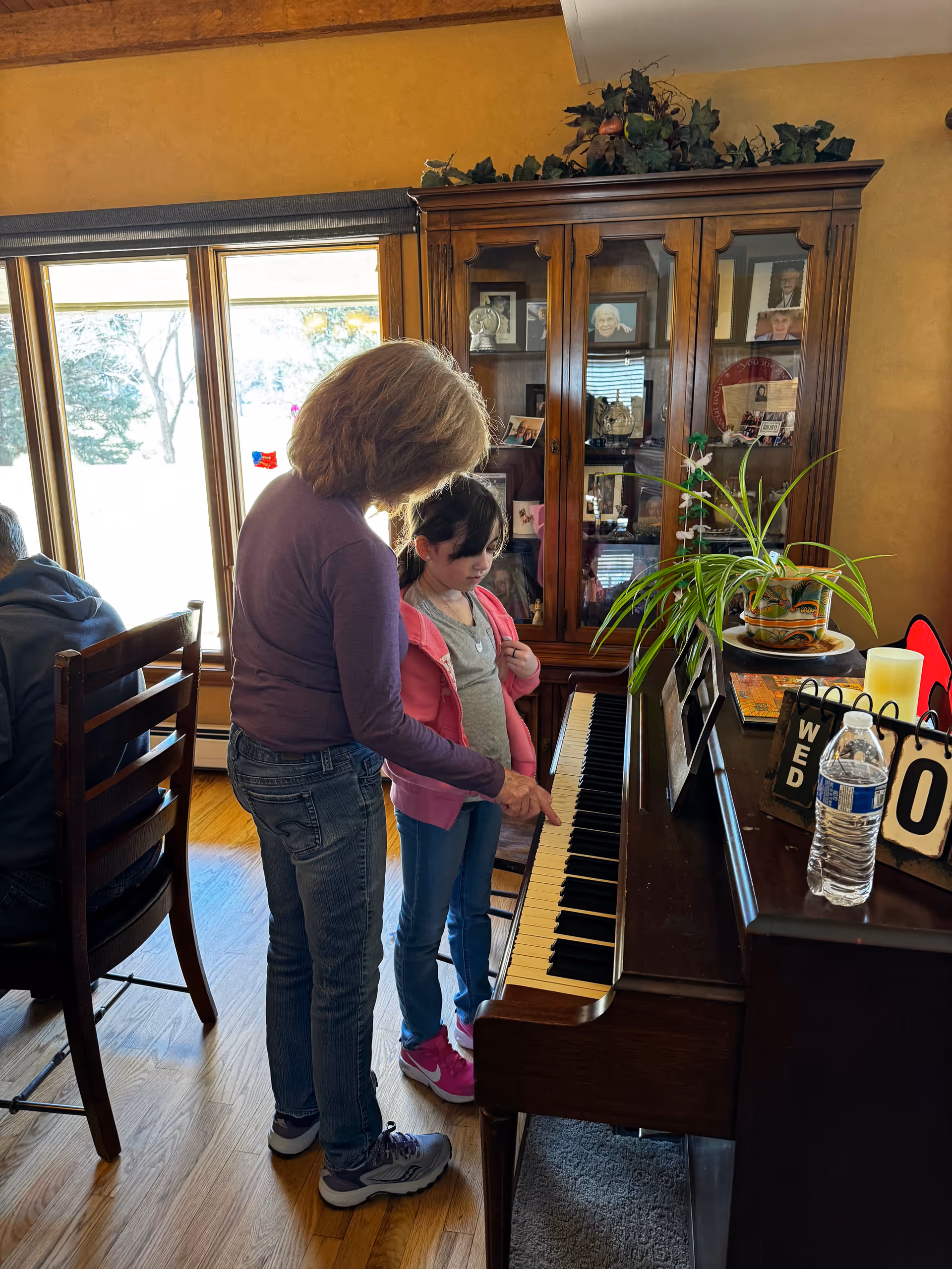 An adult woman shows a young girl how to play a piano in a cozy living room with a china cabinet and plants nearby.