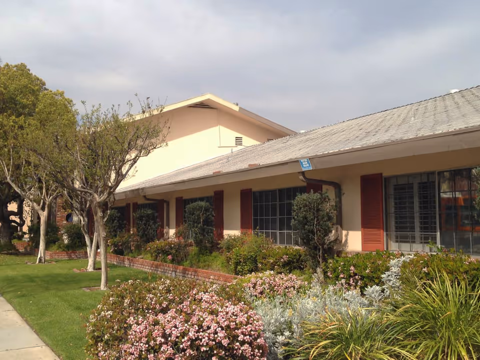 Exterior view of a single-story building with a sloped roof, large windows with red shutters, and a well-maintained garden featuring bushes, flowering plants, and small trees along a sidewalk.