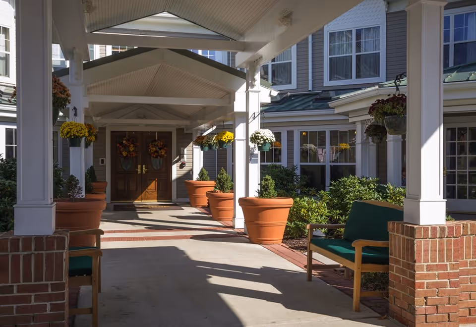 Covered entrance to a senior living facility with large potted plants and hanging flower baskets along the walkway. There are wooden double doors decorated with autumn wreaths at the end of the walkway. Green cushioned benches are placed on either side under the covered area. The building exterior is light gray with white trim and multiple windows.