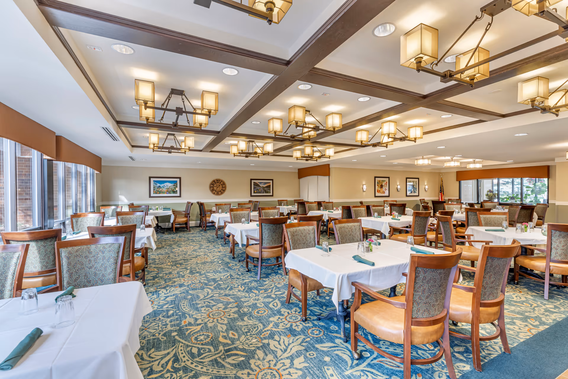 A spacious dining room with multiple tables covered in white tablecloths, each set with green napkins and glasses. The room features patterned carpet, wooden chairs with cushioned seats and backs, large windows on one side, and decorative ceiling lights.