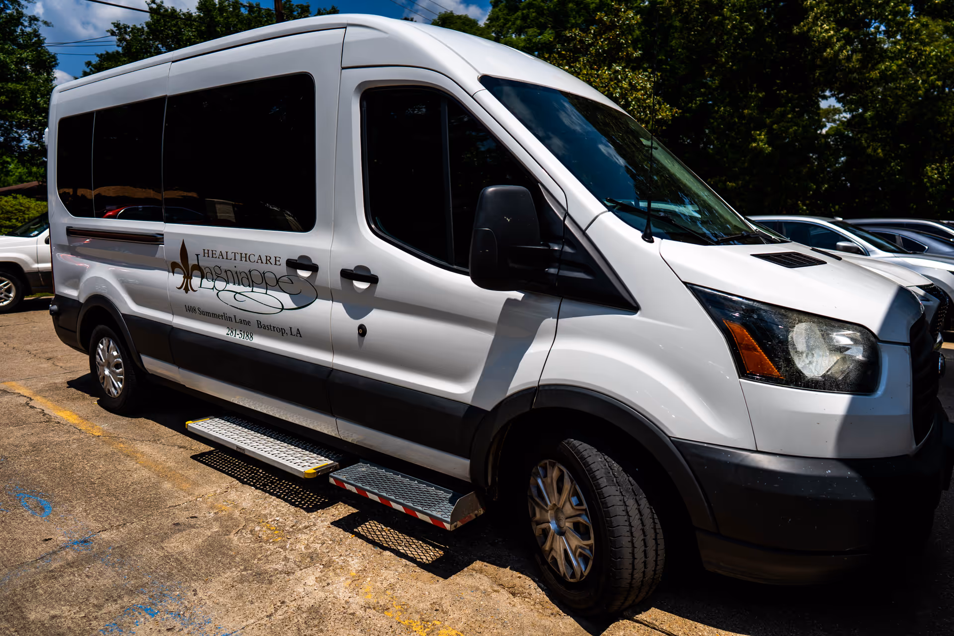 A white healthcare transport van parked in a lot with the logo and text 'Healthcare Lagniappe' on the side, along with the address 108 Summerlin Lane, Bastrop, LA and phone number 281-5188. The van has a side step for easy access and is surrounded by trees and other parked cars.