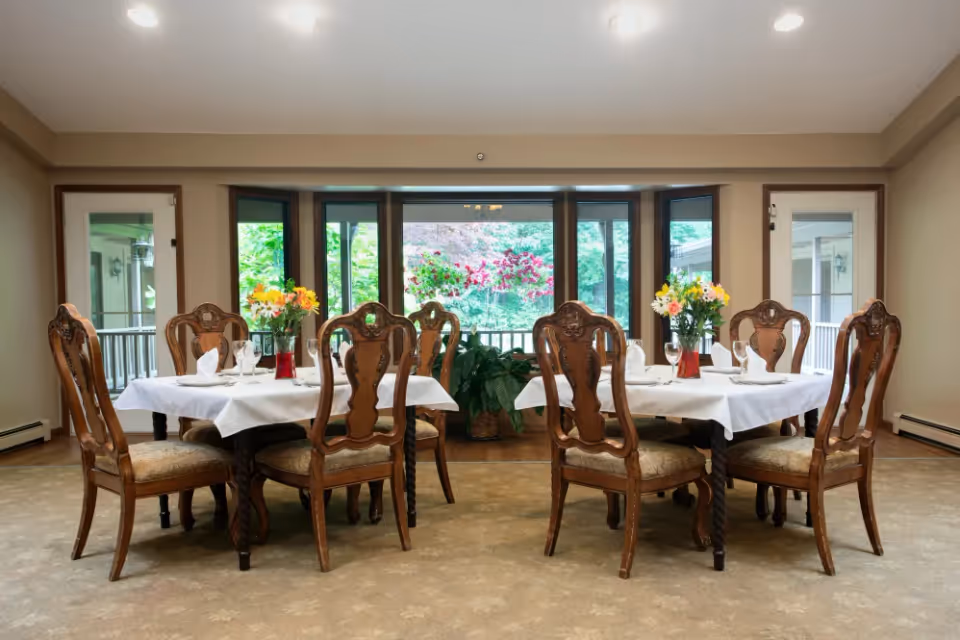 A dining room with two tables covered with white tablecloths, each surrounded by six ornate wooden chairs with cushioned seats. Each table has a vase with colorful flowers and neatly arranged glassware and napkins. Large windows and glass doors in the background reveal a green outdoor area with hanging flower baskets.
