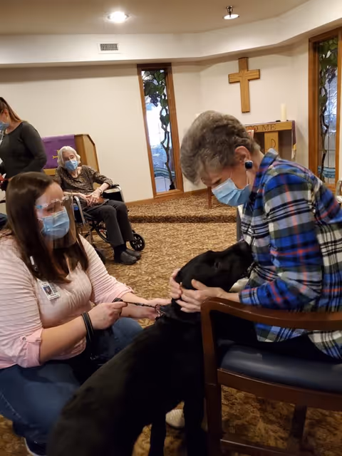 An elderly woman wearing a mask sits in a chair petting a black dog while a caregiver, also masked and wearing a face shield, kneels beside the dog holding its leash. Another elderly woman in a wheelchair is visible in the background inside a room with stained glass windows and a wooden cross on the wall.