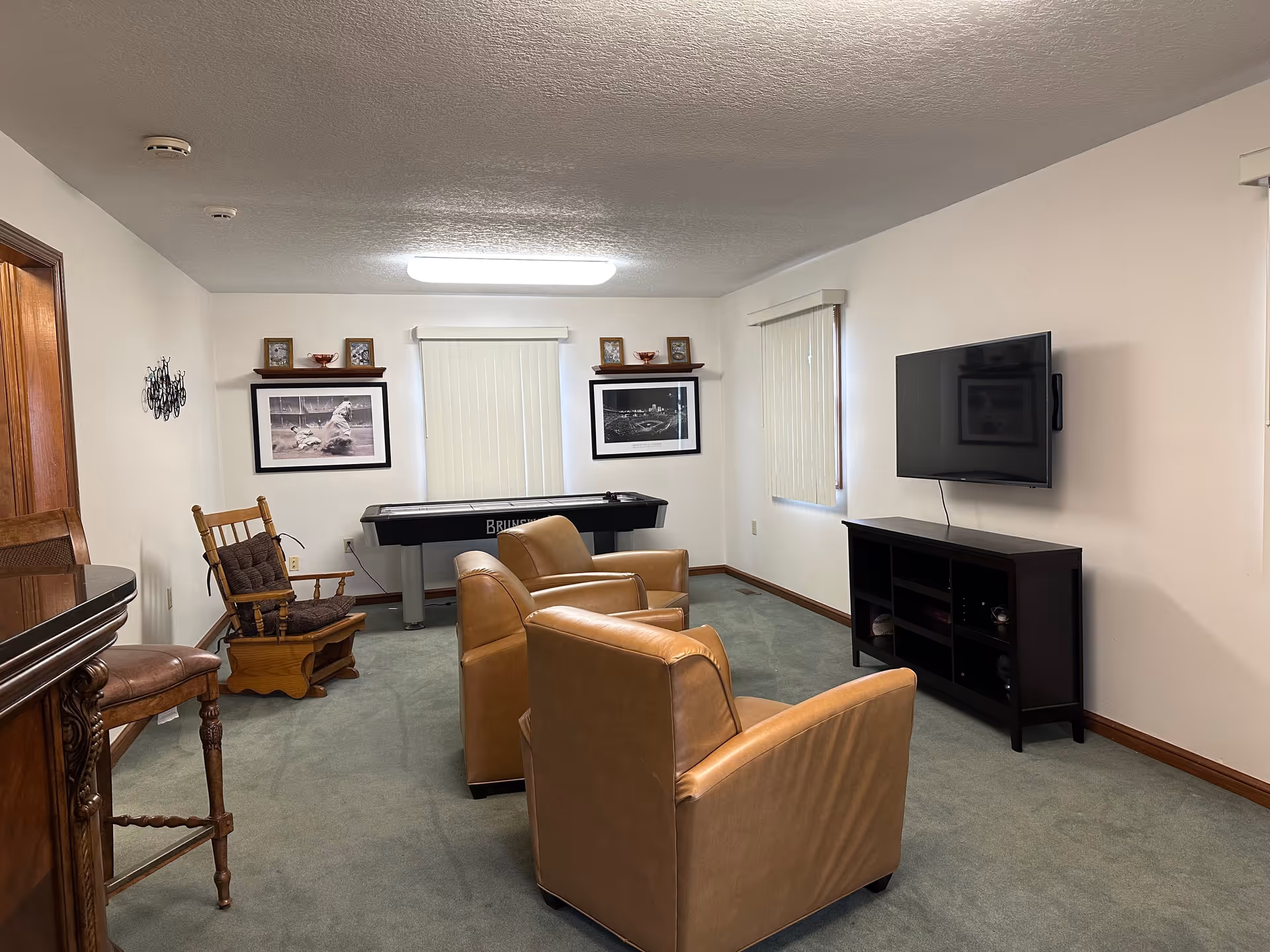 A cozy common room with two tan leather armchairs facing a wall-mounted flat screen TV above a dark wooden media console. There is a wooden rocking chair with a cushion near the left wall and a shuffleboard table against the back wall. The room has light-colored walls, green carpet, and two windows with vertical blinds. Two framed black and white photos and small decorative items are displayed on shelves above the shuffleboard table.