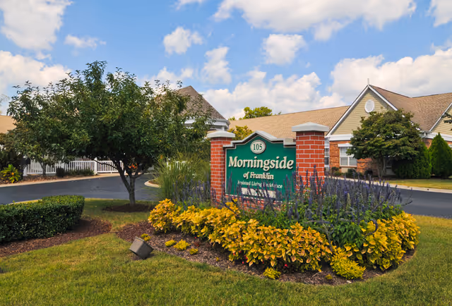 Exterior view of Morningside of Franklin assisted living residence sign surrounded by landscaping with bushes, flowers, and a tree, with the building and a partly cloudy sky in the background.