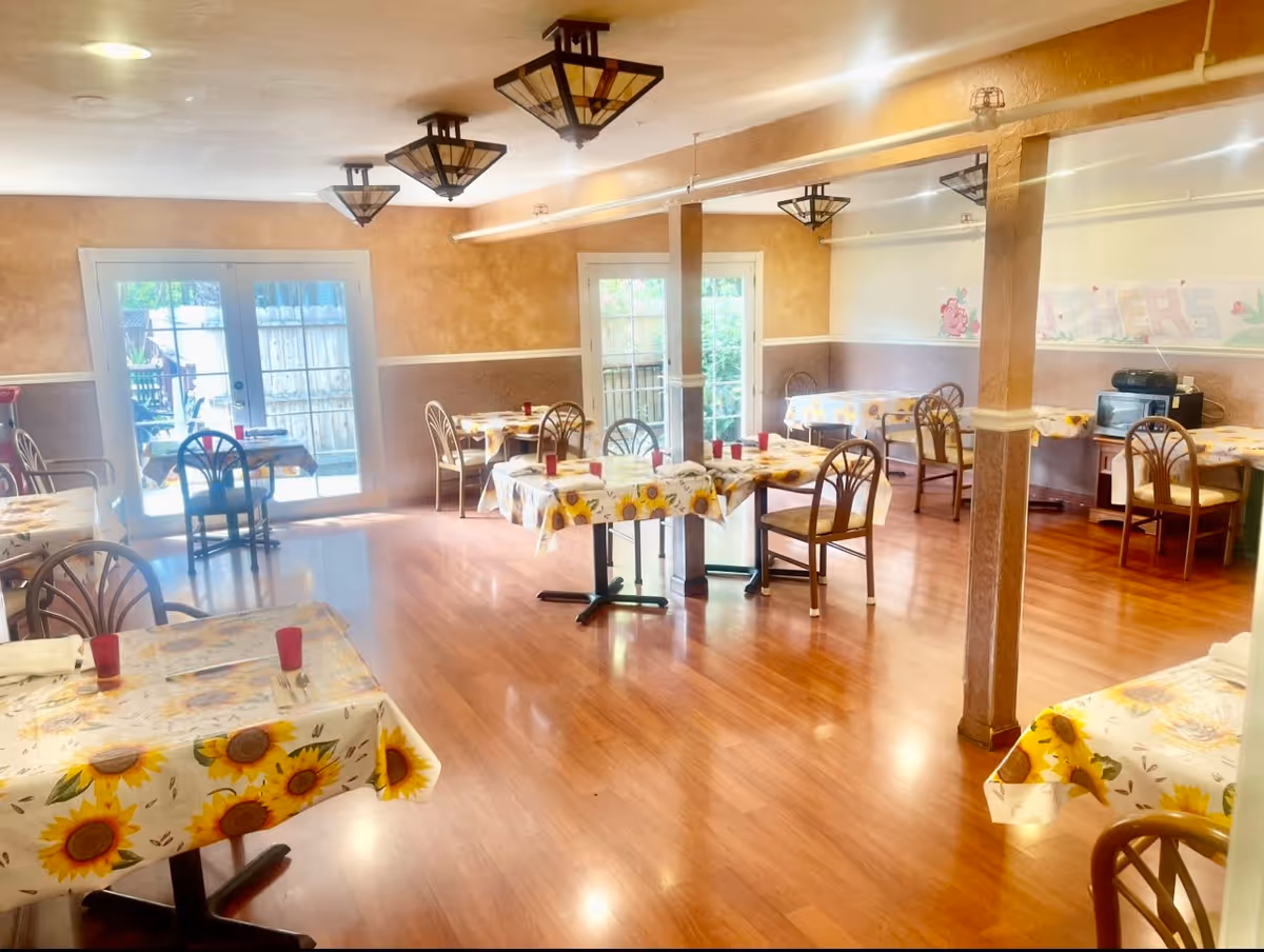 Dining room with several small tables covered in sunflower tablecloths, wooden chairs, and glass doors leading outside.