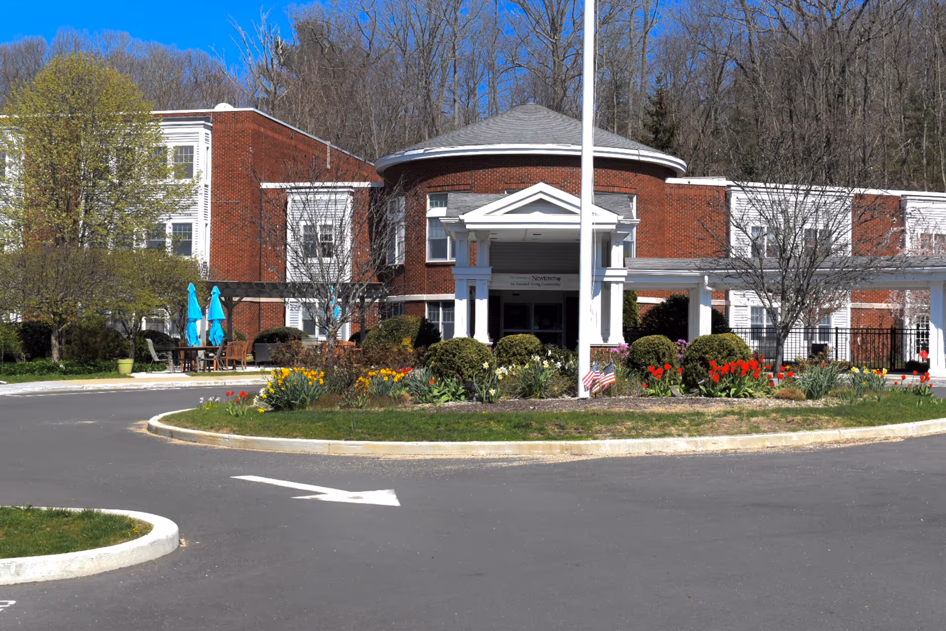 Front entrance of a red-brick senior living building with a circular portico, flagpole, landscaped flowerbeds, and a driveway roundabout.