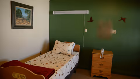 A small bedroom with a single bed, patterned bedding, a wooden nightstand, and framed wall art on a green accent wall.