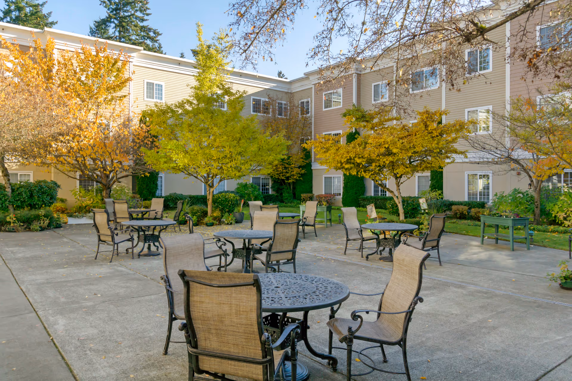 Outdoor courtyard area of a senior living facility with several round metal tables and cushioned chairs arranged on a concrete patio. The courtyard is surrounded by a three-story beige building with white trim and multiple windows. Trees with autumn-colored leaves and green shrubs add greenery to the space under a clear blue sky.