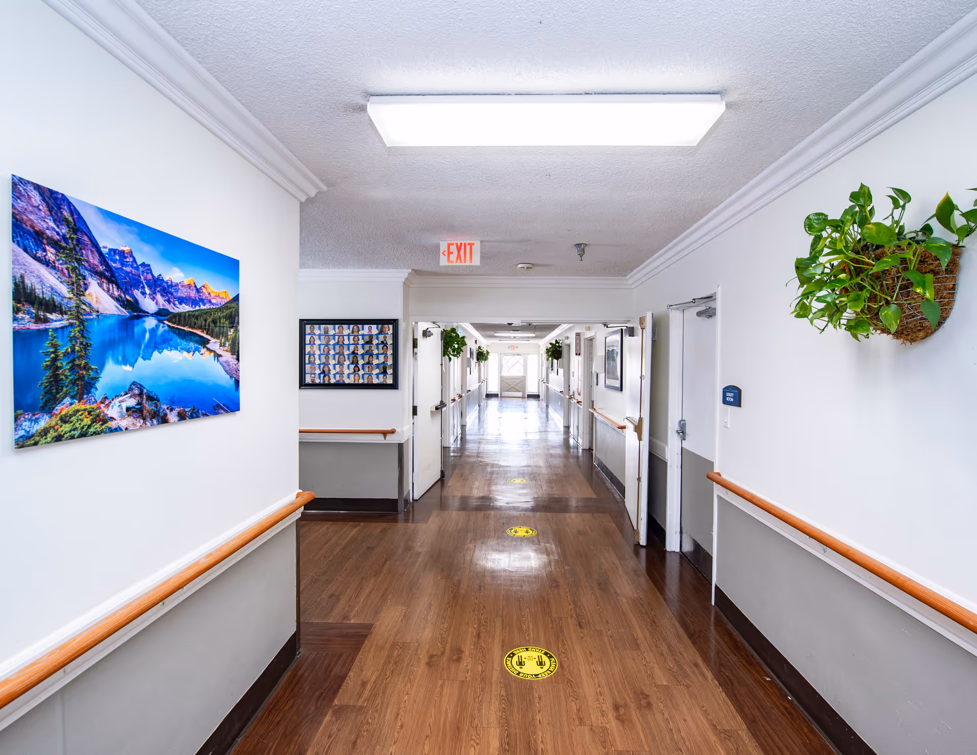 A clean, well-lit hallway in Pomona Vista Care Center with wooden flooring, white walls, handrails on both sides, a large landscape photograph on the left wall, a framed collage of portraits further down the hall, and green plants mounted on the right wall. An exit sign is visible on the ceiling near the end of the hallway.