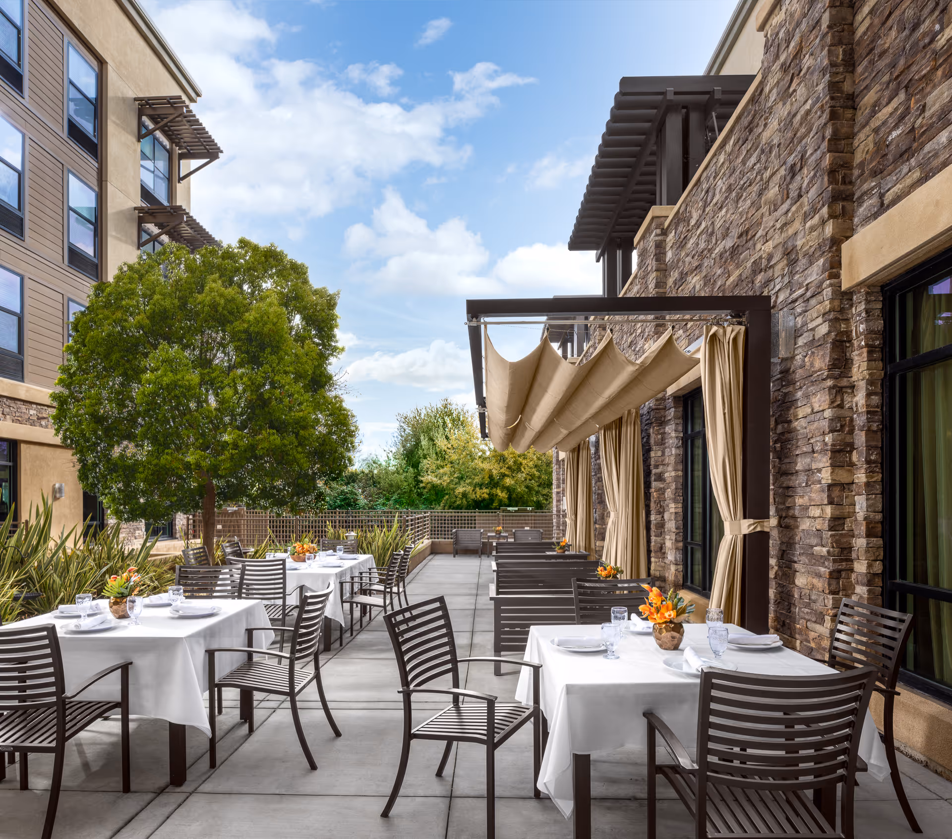 Outdoor patio dining area at Belmont Village Senior Living Albany with tables covered in white tablecloths, brown metal chairs, small flower arrangements on each table, a large green tree, and a stone building wall with beige curtains and a retractable awning under a partly cloudy sky.