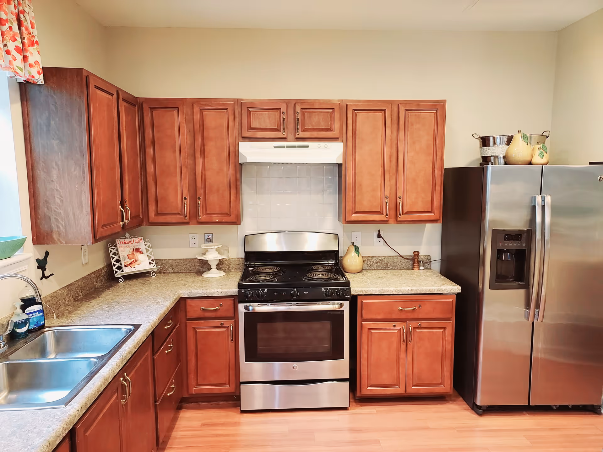 A kitchen with wooden cabinets, a stainless steel electric stove with oven, a stainless steel refrigerator with water and ice dispenser, a double sink, and a countertop with decorative items including a cookbook on a stand and ceramic pitchers.