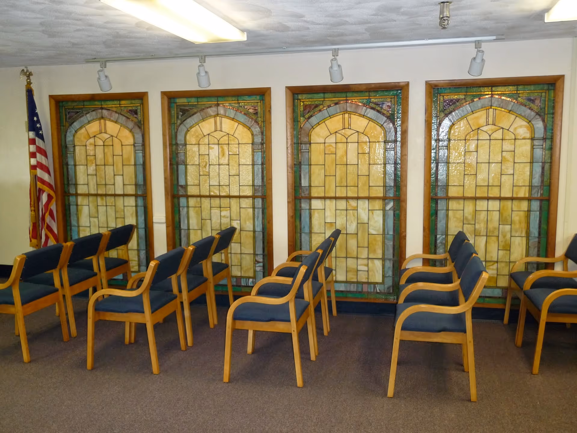 Interior room with rows of wooden chairs with blue cushions arranged facing stained glass windows. An American flag is positioned in the corner of the room.