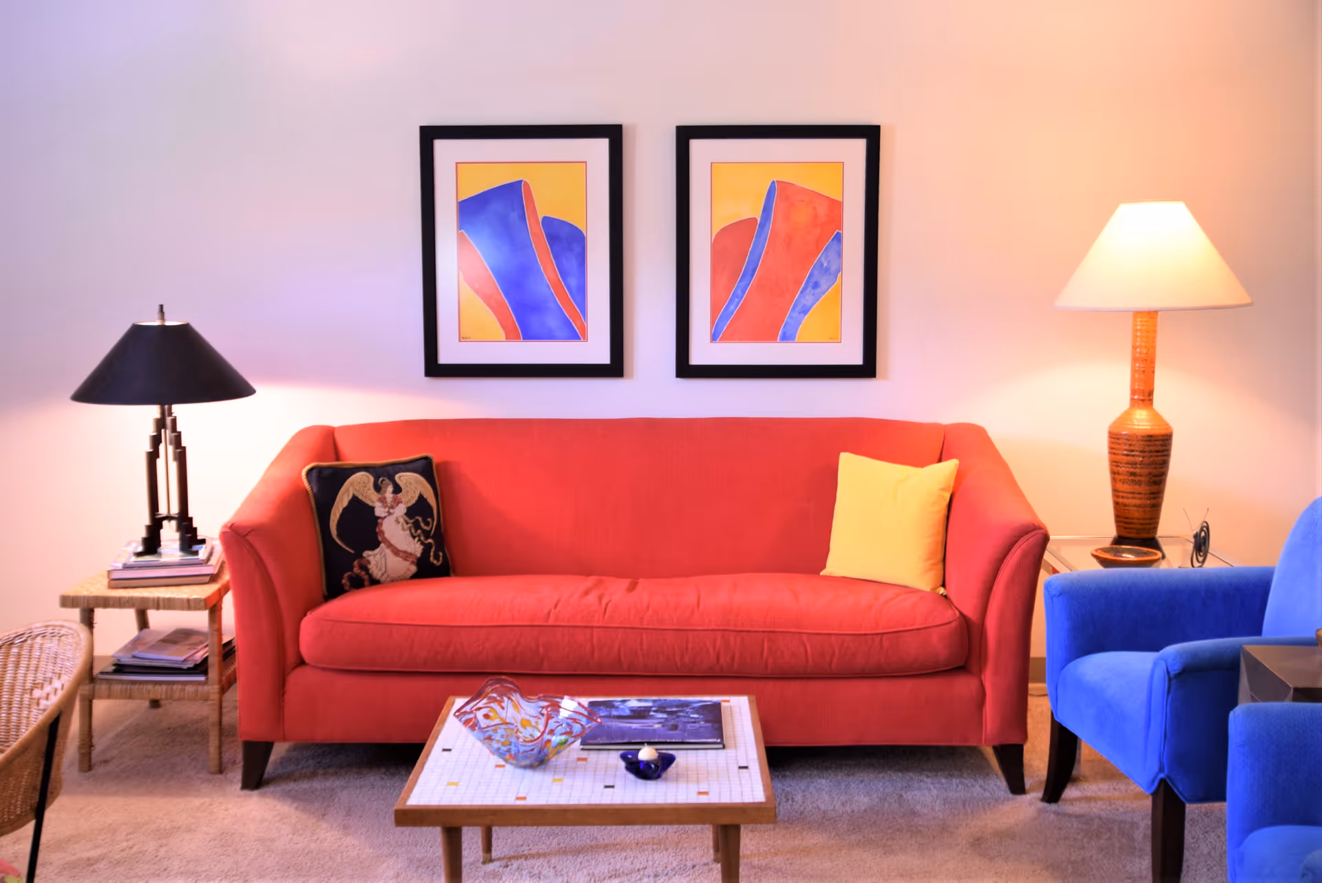 Red sofa with a yellow pillow centered beneath two colorful framed prints, flanked by table lamps, a coffee table, and a blue armchair.