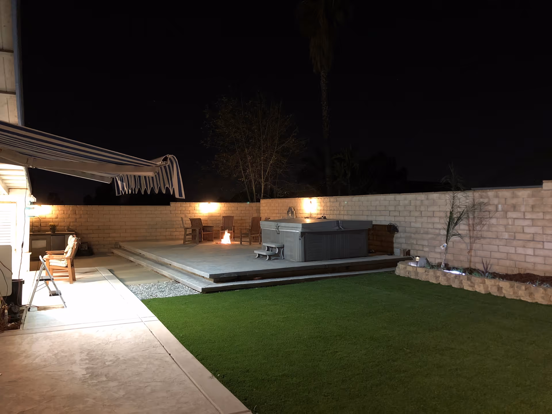 Backyard patio at night showing a raised deck with a hot tub, fire pit, seating and a lawn.