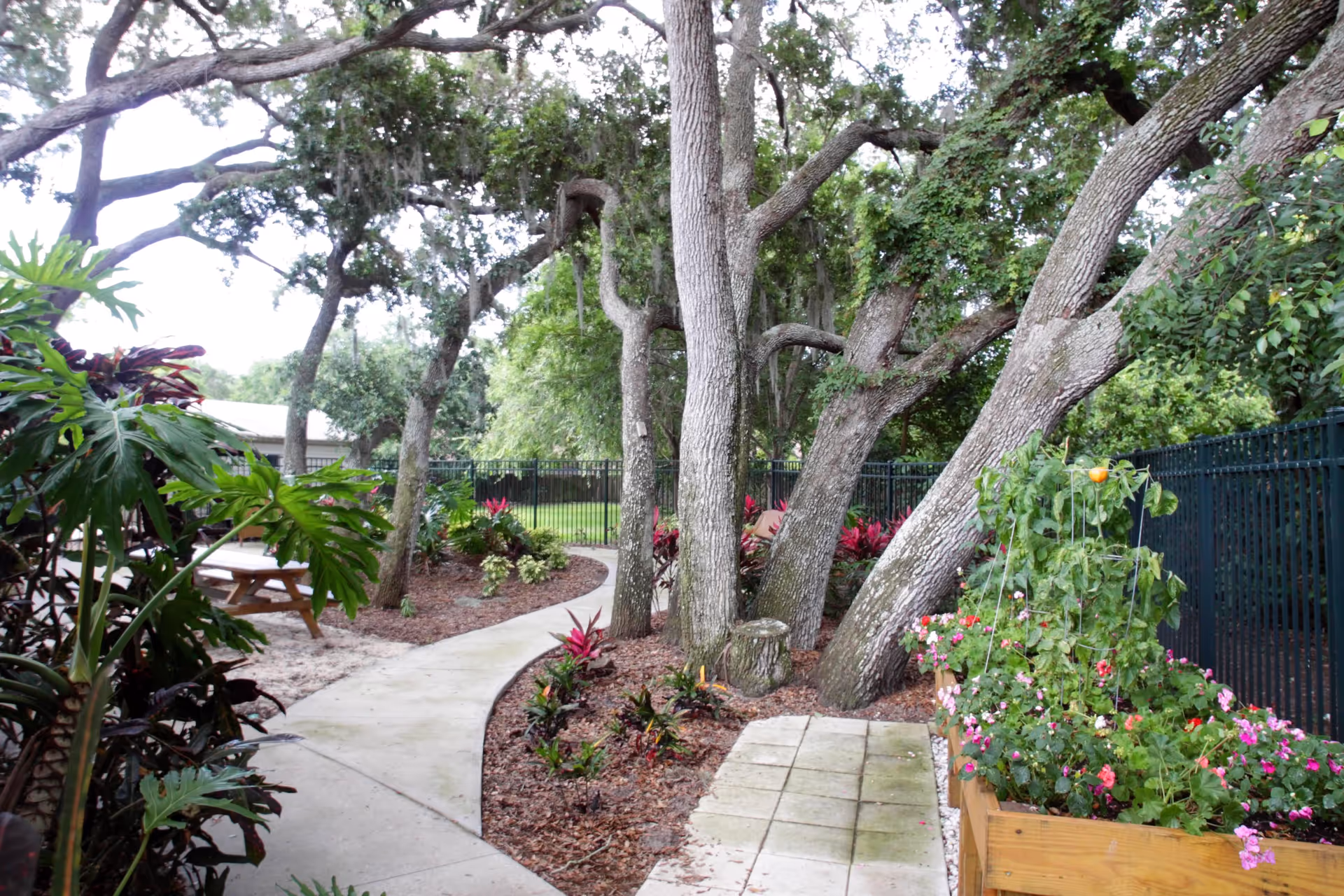 A peaceful outdoor garden area with a curved concrete walkway surrounded by trees, plants, and flowers. There is a wooden picnic table on the left side and a raised wooden planter box with flowering plants on the right. A black metal fence encloses the area.