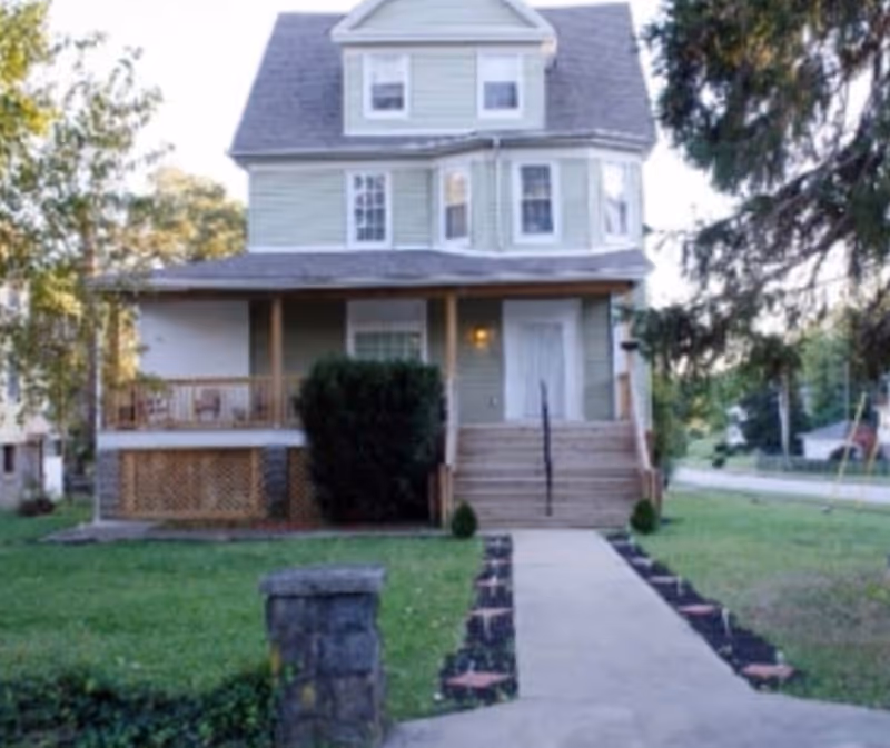 Front exterior view of a two-story light green house with a porch, stairs leading up to the front door, and a walkway lined with small plants. The house is surrounded by grass and trees.