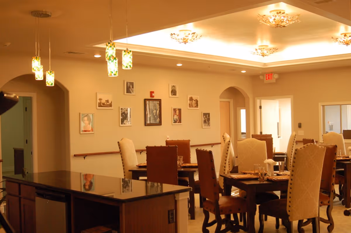 A warm, well-lit dining room with several wooden tables and upholstered chairs arranged neatly. The room features pendant lights with green and yellow stained glass shades hanging over a dark granite countertop island. The walls are decorated with framed black and white and color photographs. There are arched doorways and an exit sign above one door.