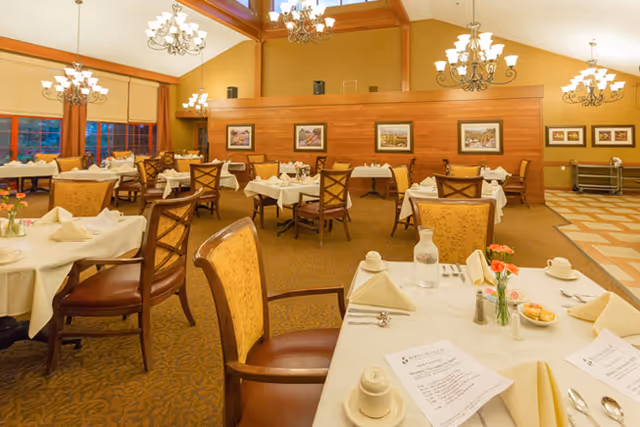 A spacious dining room with multiple round tables covered with white tablecloths, set with napkins, menus, cups, and silverware. The room features warm yellow walls, wooden accents, chandeliers hanging from a high ceiling, and framed artwork on the walls. Chairs with cushioned seats and backs surround the tables.