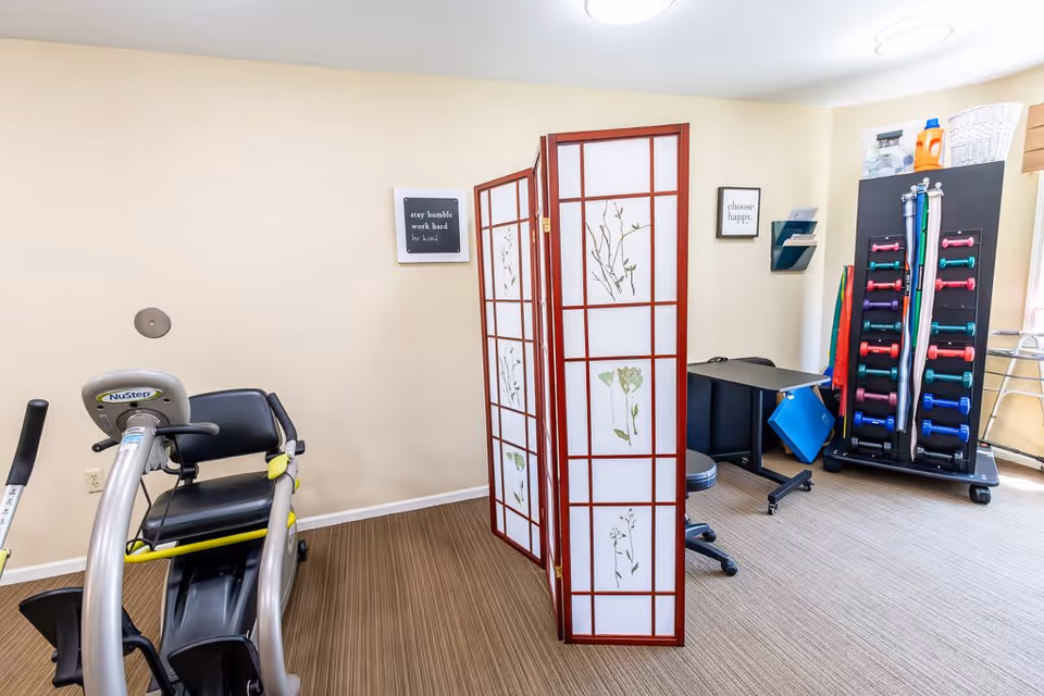 A small exercise room with a NuStep recumbent cross trainer, a decorative folding screen with floral designs, a desk with a rolling chair, and a rack holding colorful dumbbells. The walls are light beige with two framed motivational quotes: 'stay humble work hard be kind' and 'choose happy.'