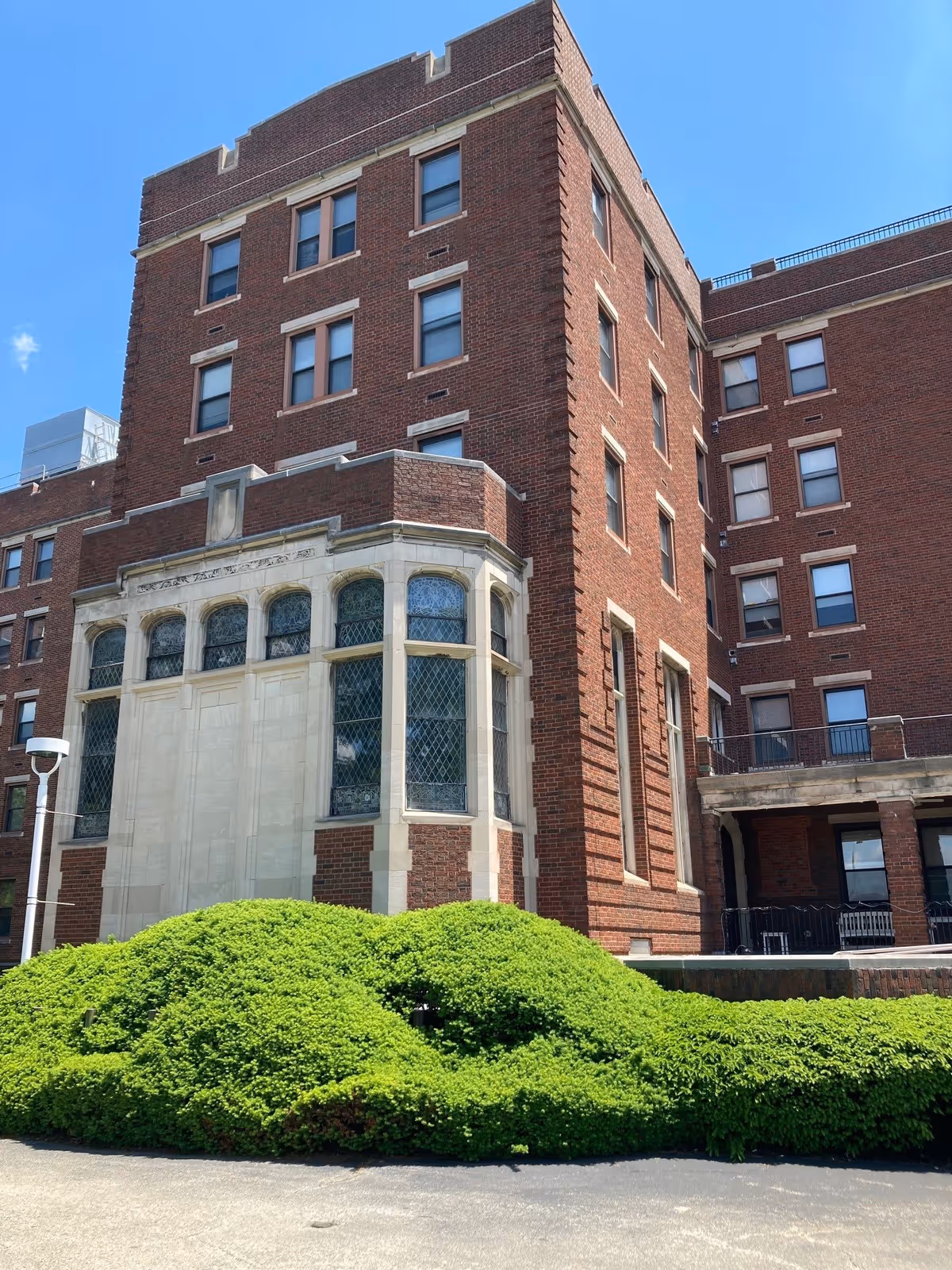 Exterior view of a multi-story brick building with large windows and decorative stonework. In front of the building, there is a well-maintained green shrubbery under a clear blue sky.