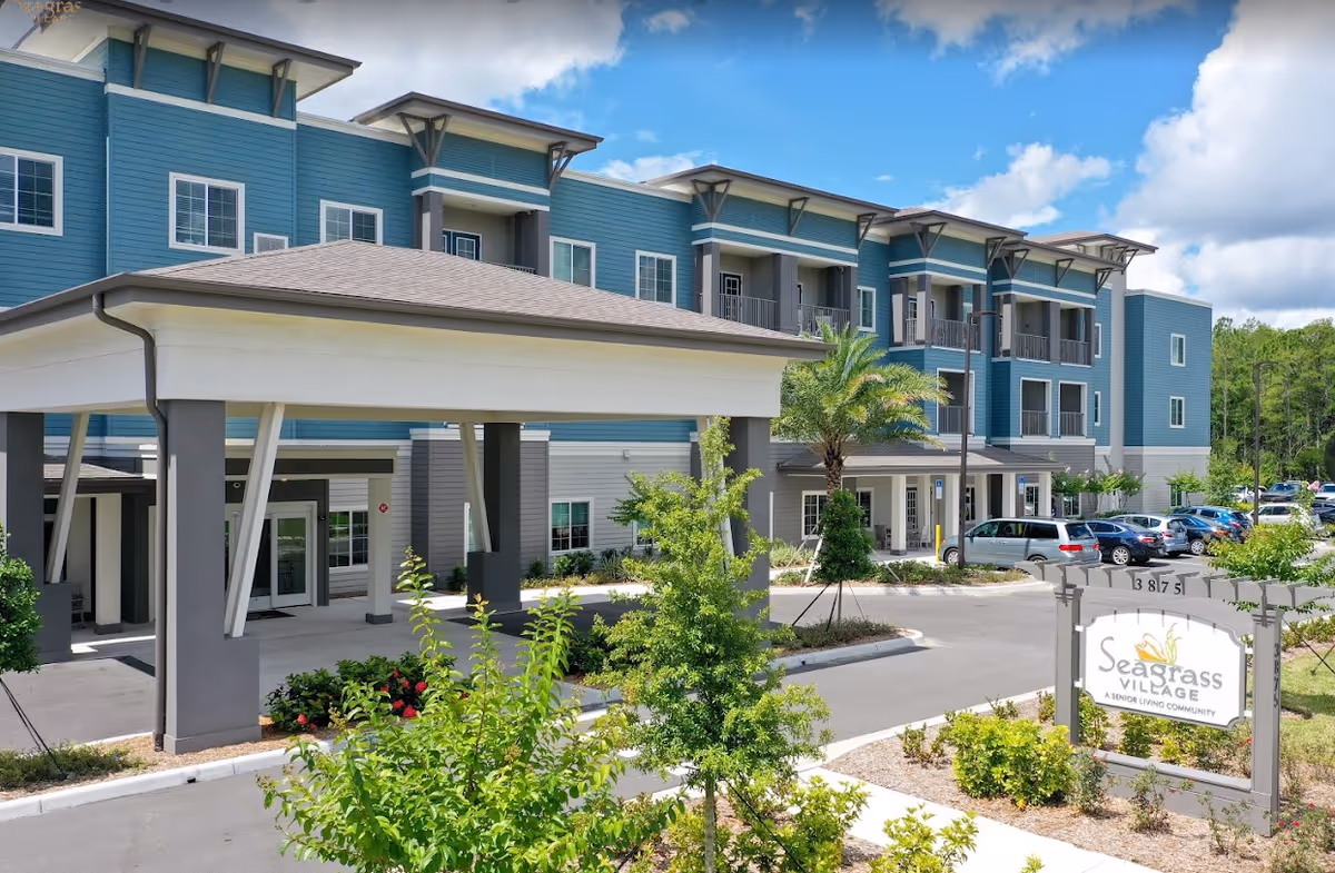 Exterior view of Seagrass Village of Port Orange senior living community building with a covered entrance, parking lot, landscaped greenery, and a sign displaying the facility name.