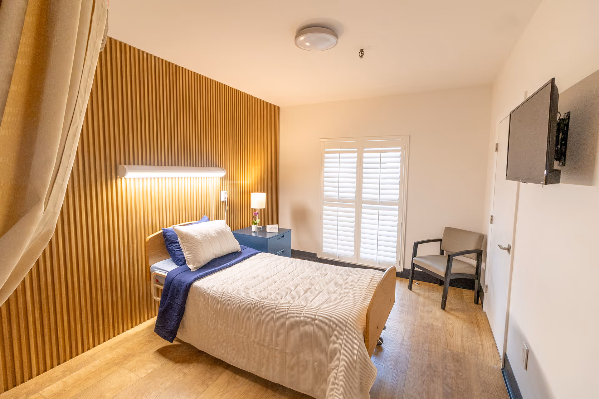 A clean and well-lit bedroom in a senior living facility with a single bed made with white and blue bedding, a wooden slatted accent wall behind the bed, a small blue nightstand with a lamp and a small flower vase, a window with white shutters, a gray armchair, and a wall-mounted TV.