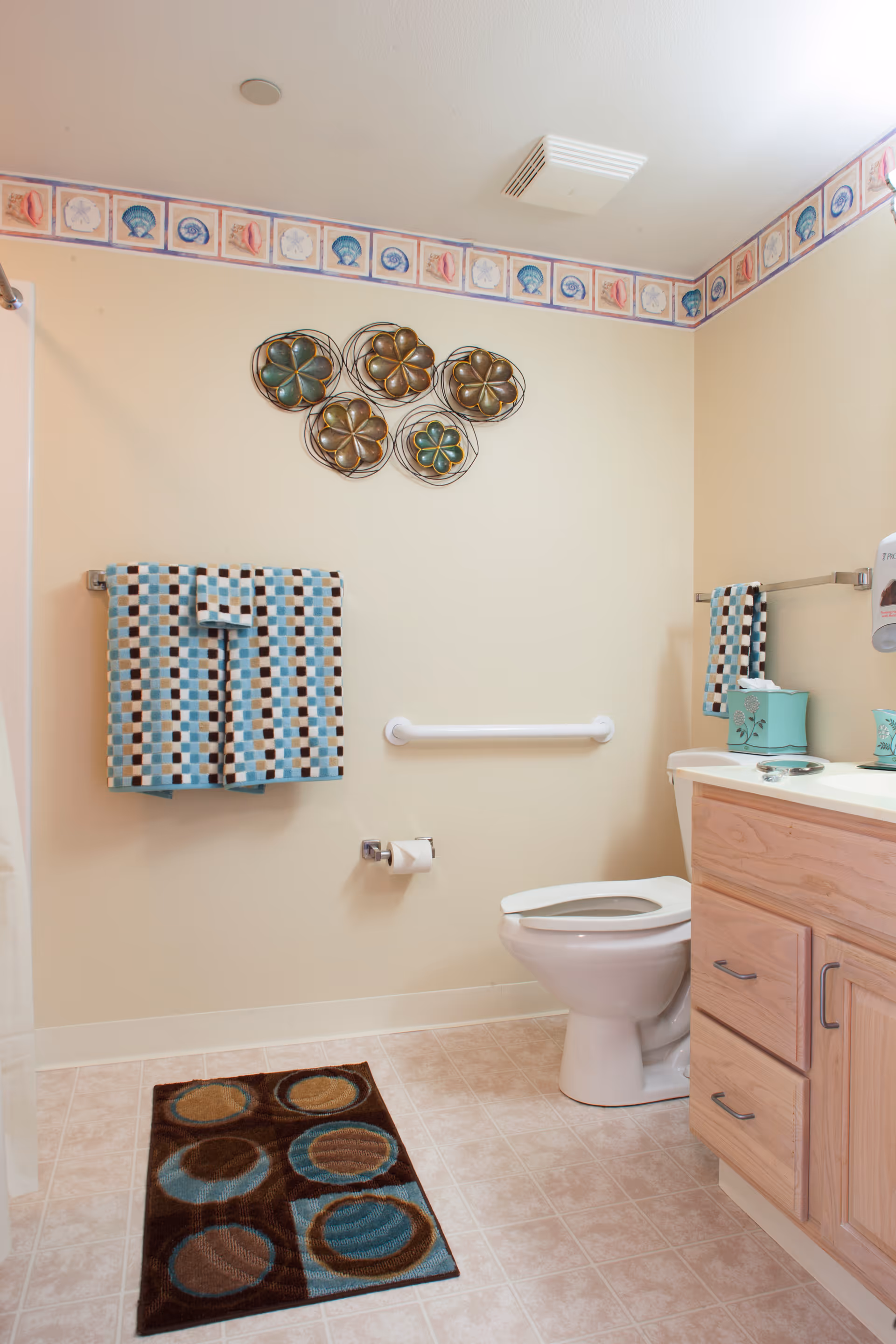 Well-lit bathroom with a toilet, wooden vanity, patterned towels and rug, decorative wall art and a safety grab bar.