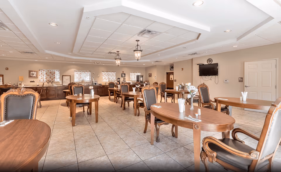 A spacious dining room with multiple wooden tables and cushioned chairs arranged neatly. Each table has utensils and napkins set, with some tables featuring small flower vases. The room has tiled flooring, beige walls, and a ceiling with recessed lighting and hanging lantern-style light fixtures. Windows with floral curtains are visible in the background, along with a wall-mounted TV and a clock.