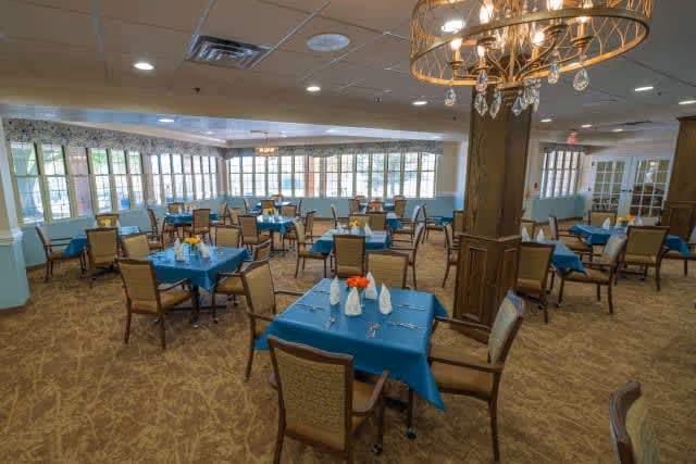 A spacious dining room with multiple tables covered in blue tablecloths, each set with white napkins, silverware, and small flower centerpieces. The room has large windows allowing natural light, beige patterned carpet, and a decorative chandelier hanging from the ceiling.