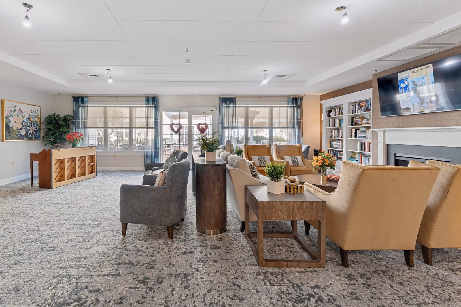 A spacious and well-lit living room area in a senior living facility with multiple comfortable armchairs and sofas arranged around wooden side tables. There is a large flat-screen TV mounted above a fireplace on the right side, and built-in bookshelves filled with books next to it. Large windows with sheer curtains allow natural light to fill the room. A piano and a large floral painting are visible on the left side of the room.