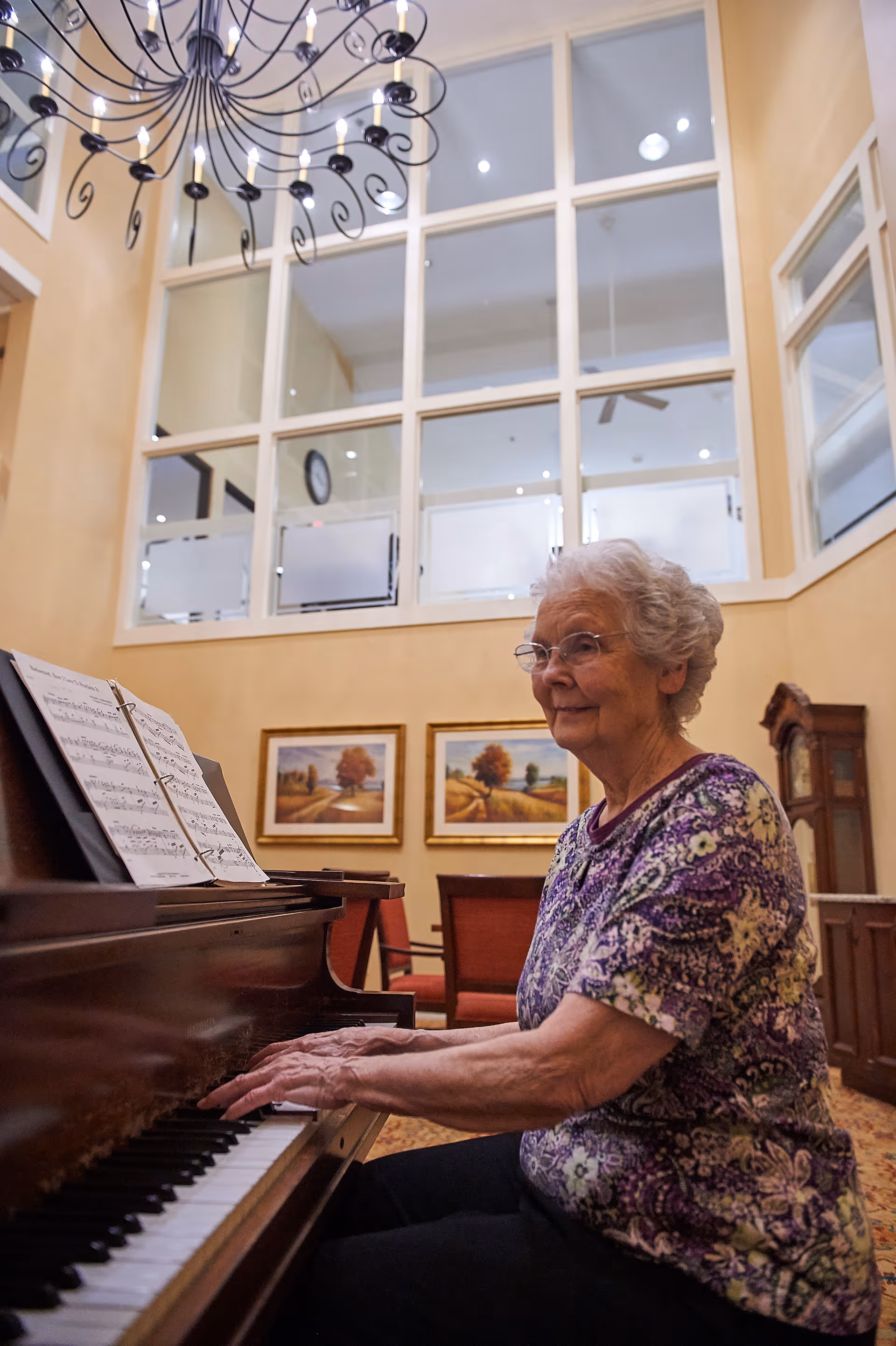 An elderly woman with glasses and short gray hair is playing a piano in a well-lit room with high ceilings and large windows. The room features a decorative chandelier, framed landscape paintings on the wall, and comfortable chairs in the background.