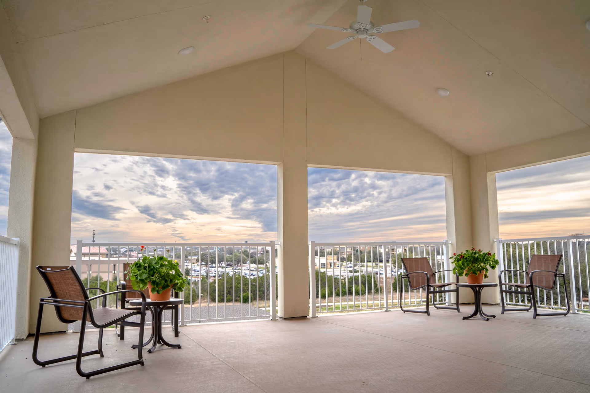 Covered outdoor patio area with a high vaulted ceiling and ceiling fan. The space has two sets of brown chairs with small tables, each table holding a potted plant. The patio overlooks a scenic view of trees, buildings, and a cloudy sky at sunset.