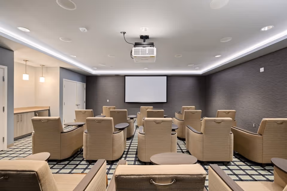 A modern media room with beige recliner chairs arranged in rows facing a large white projection screen on a dark textured wall. The ceiling has recessed lighting and a mounted projector. The floor is carpeted with a patterned design, and there is a small counter area with pendant lights on the left side of the room.