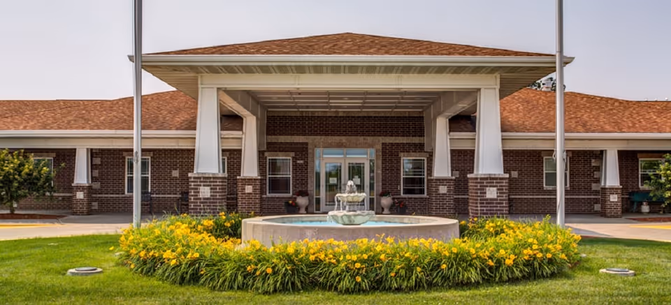 Front entrance of a brick senior living building with a covered portico, central fountain and a ring of yellow flowers.