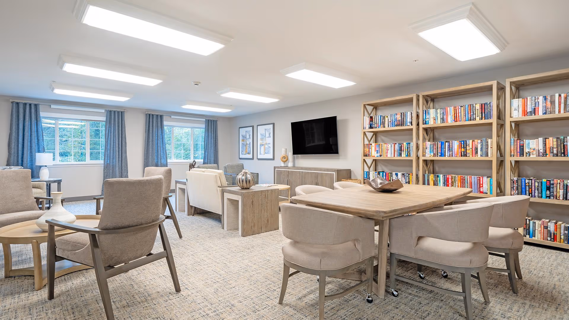 A bright and spacious common area in a senior living facility featuring multiple comfortable beige armchairs and sofas arranged around wooden tables. The room has large windows with blue curtains letting in natural light, a wall-mounted flat-screen TV, and several wooden bookshelves filled with books along one wall. The carpet is light-colored with a subtle pattern, and the ceiling has recessed lighting panels.