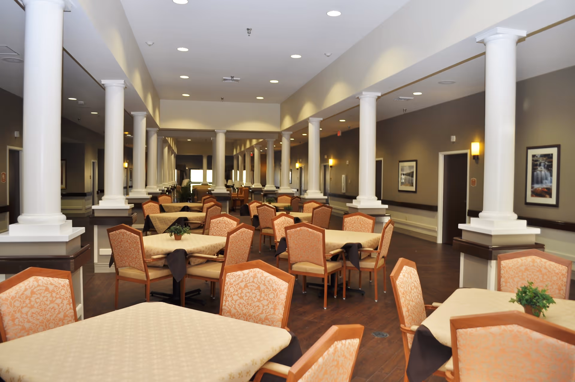 A spacious dining room with multiple tables covered with beige tablecloths and surrounded by cushioned chairs with patterned upholstery. The room features white columns, warm lighting, framed pictures on the walls, and a wooden floor.