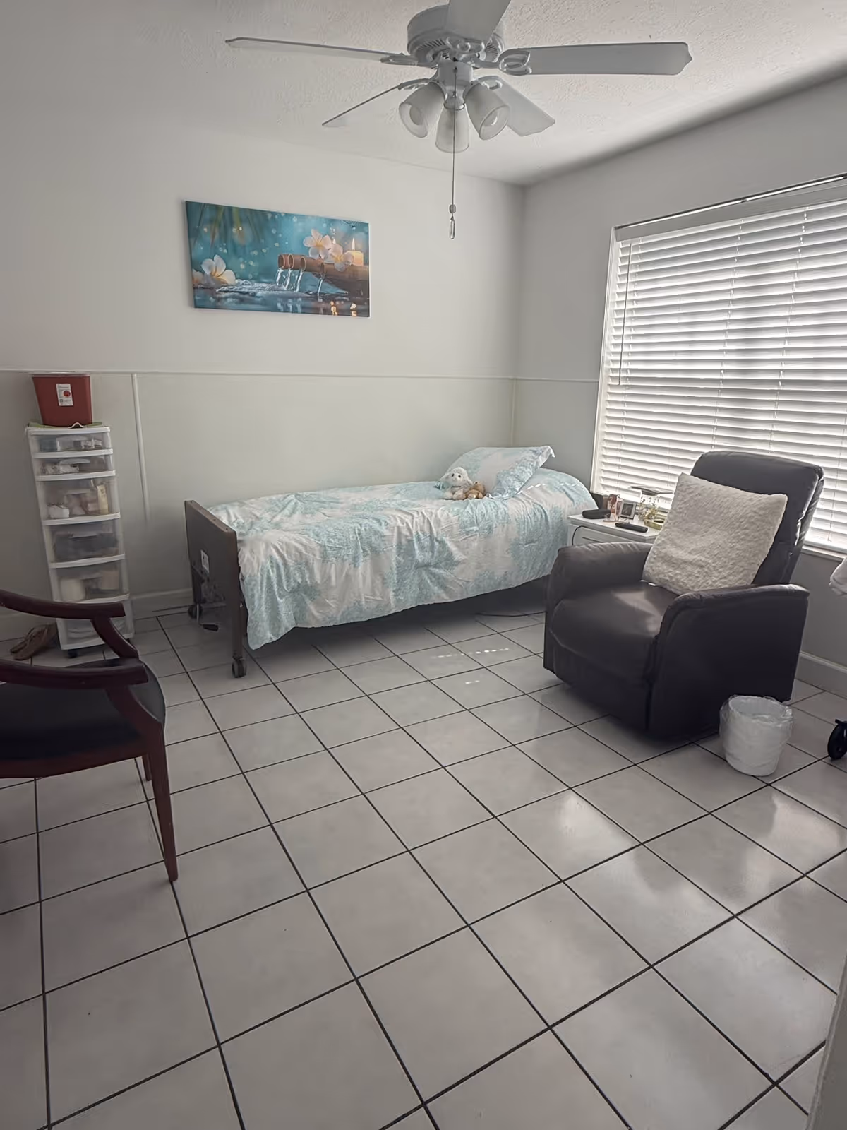 A small, simple bedroom with a single bed covered in a light blue and white patterned bedspread. There is a stuffed animal on the bed. Next to the bed is a small white nightstand with various items on it. A large window with closed blinds is on the right wall. A dark recliner chair with a white pillow is positioned near the window. On the left side of the room, there is a wooden chair and a plastic drawer storage unit. The floor is tiled with white tiles and the walls are painted white. A ceiling fan with lights is mounted on the ceiling. A decorative painting featuring water and flowers hangs above the bed.