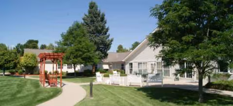 Exterior view of Brookdale Pinehurst Park facility with green lawn, trees, and a red arbor.