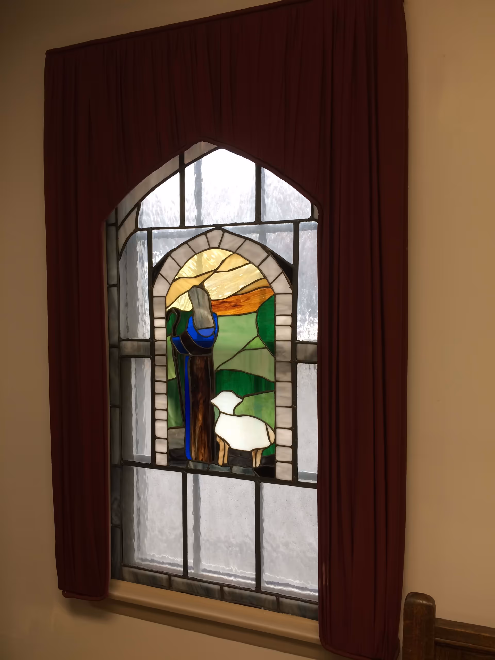 A stained glass window with a religious design depicting a figure in blue and brown robes standing next to a white lamb, set against a background of green hills and a yellow sky. The window is framed by maroon curtains and is set in a beige wall.
